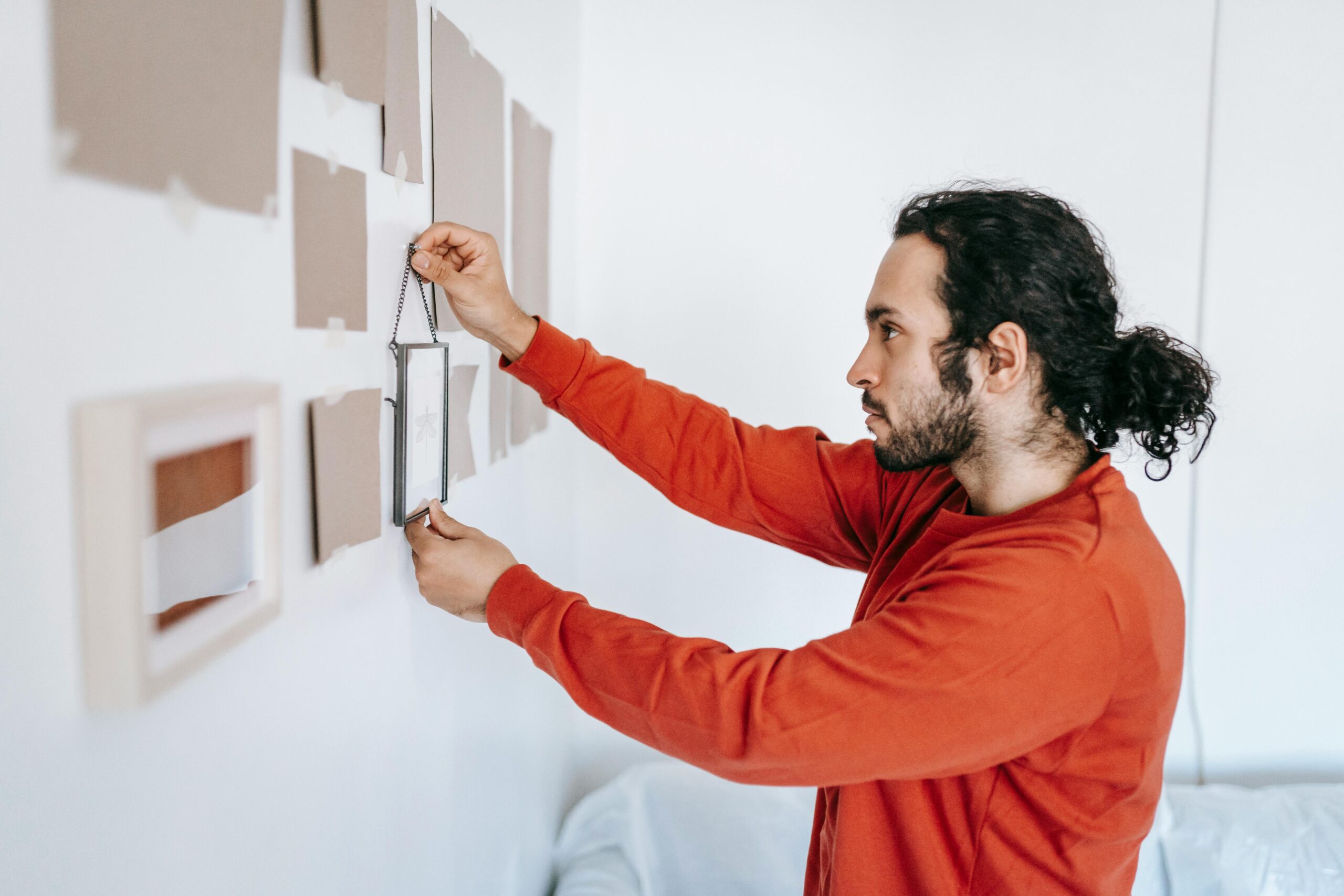 A man in a red sweater hangs a picture frame on a white wall in a modern room.